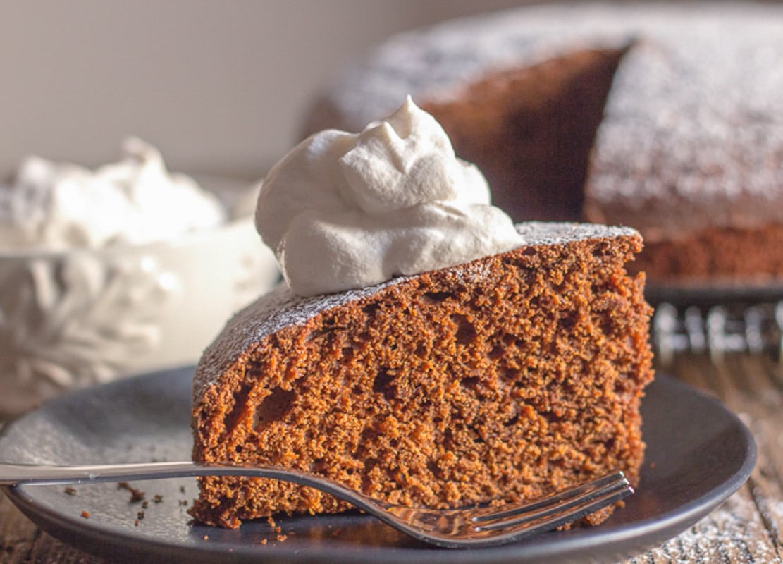 Gingerbread cake on a wire rack with a slice with a dollop of whipped cream on a black plate.