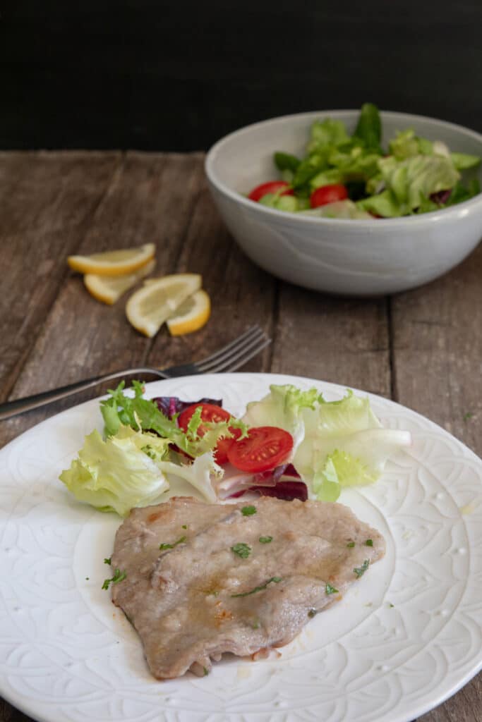 A slice of veal on a white plate with some salad on the plate and in a bowl.