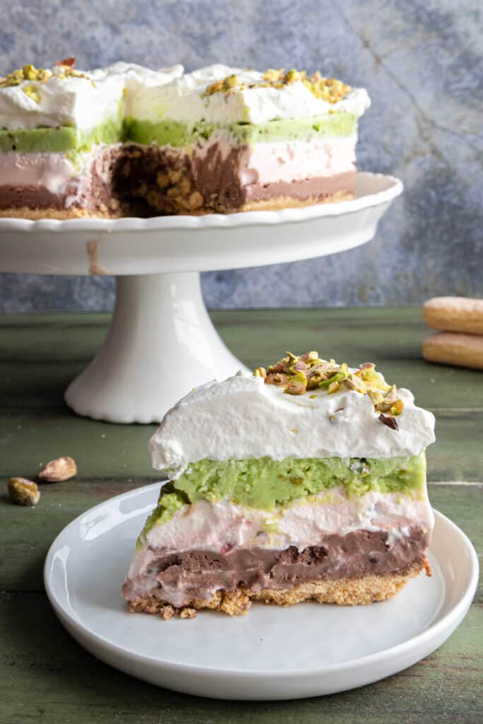 Ice cream cake on a cake stand with a slice on a white plate.