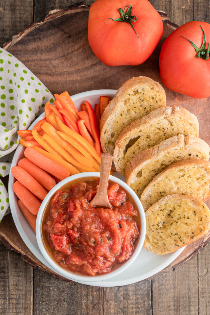 Tomato dip in a white bowl with crackers and carrot sticks.