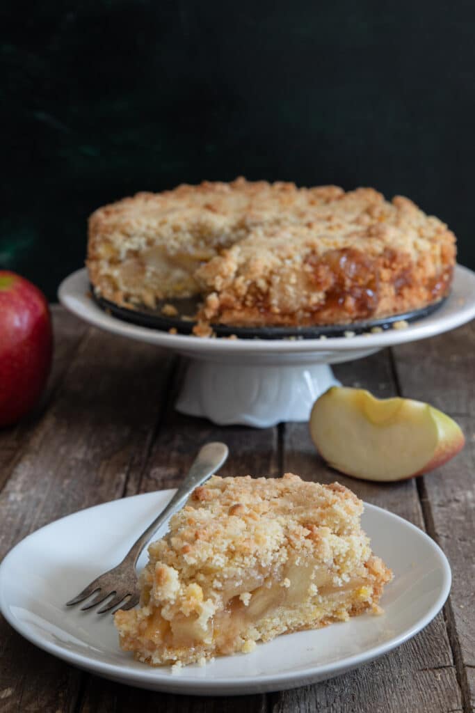 Apple crumb cake on a white cake stand with a slice on a white plate.