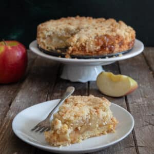 Apple crumb cake on a white cake stand with a slice on a white plate.