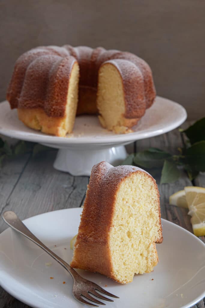 Lemon cake on a white cake stand and a slice with a fork on a small white plate.