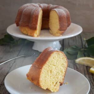 Lemon cake on a white cake stand and a slice with a fork on a small white plate.