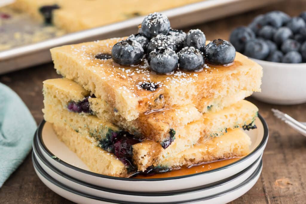 Four pancakes packed on a white plate with blueberries and syrup.