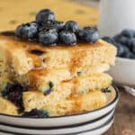 Four pancakes packed on a white plate with blueberries and syrup.