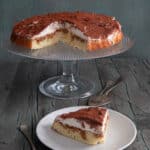 Tiramisu crostata on a glass cake stand with a slice on a white plate.