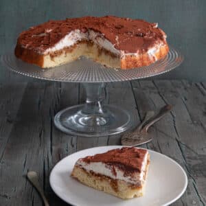 Tiramisu crostata on a glass cake stand with a slice on a white plate.
